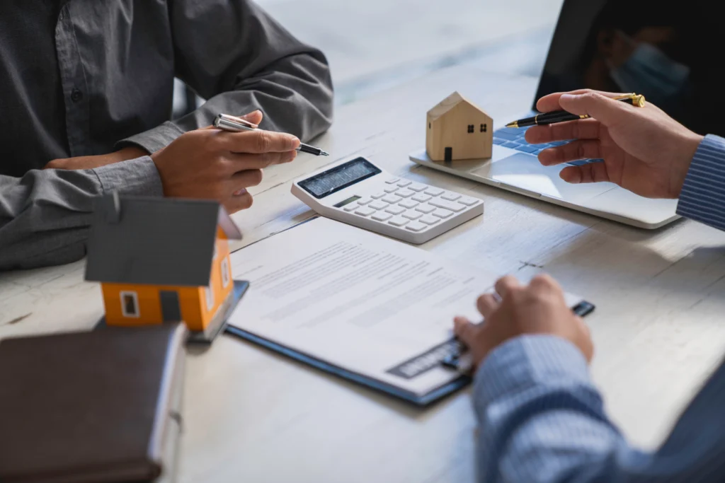 People signing paperwork beside model houses and a calculator